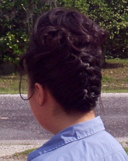 Close up of the back of Elysia's hair style, a frech braid that starts at the nape of the neck and ends in soft curls at the top back of her head. Elysia is standing outside, with grass, road and bushes just visible in the background. (75974 bytes)