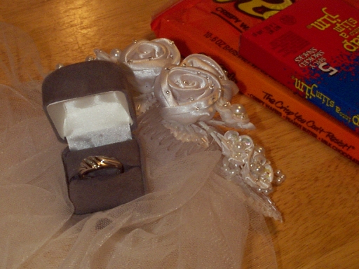 Close up of an open grey velvet ring box with man's ring inside, box on veil with beaded head piece, the corner of packages of chocolate bars showing in the background.(148541 bytes)