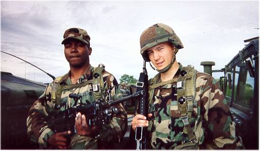 A picture of two young men in combat fatigues with helmets on.  On the Left is Scott's training buddy, and to the right is Scott. Both are holding wicked looking guns.