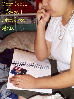 wide shot of photo above showing the hand, picture and the entire notebook, pen in right hand, showing part of Elysia profile of Elysia's face from the nose down, and body sitting on bed cross legged with notebook on lap, face from nose down.  Text in curly handwriting font on image: Dear Scott, Today I missed you again. A silver chain with a man's class ring hangs around her neck.